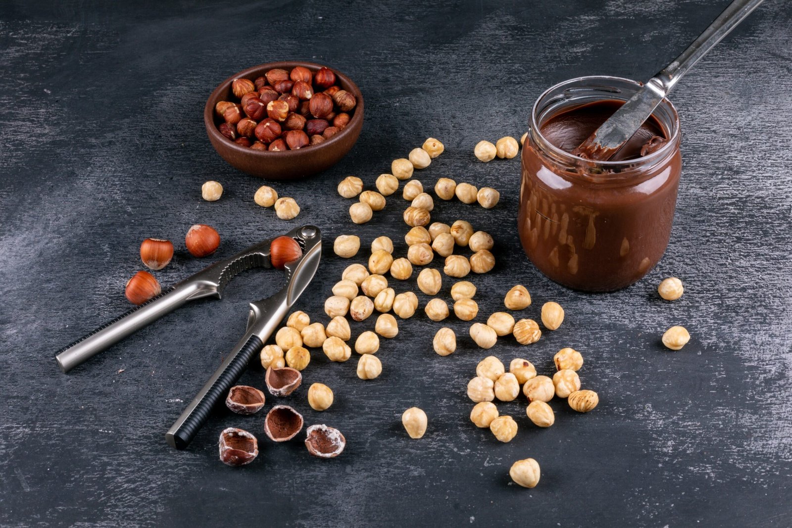 high-angle-view-scattered-cleaned-hazelnuts-brown-bowl-with-cocoa-spread-nutcracker-dark-stone-table-horizontal