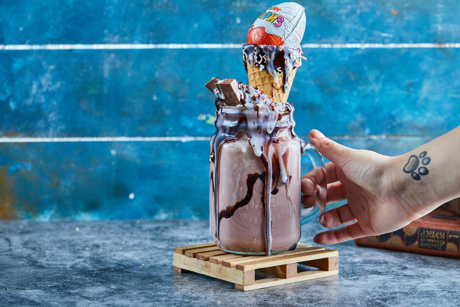 woman-hand-with-tattoo-holding-mug-chocolate-smoothie-with-kinder-surprise-sprinkles-blue-surface